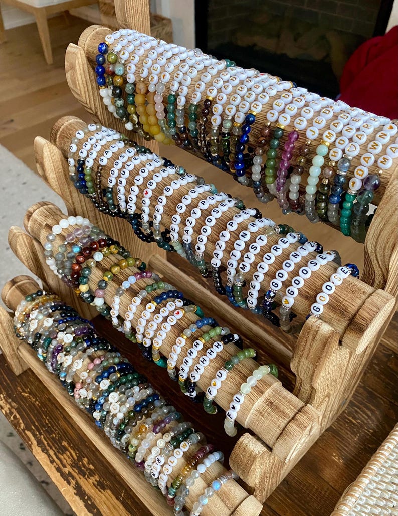 May include: A wooden display rack holds numerous beaded bracelets. The bracelets feature a variety of colorful beads and white letter beads spelling out words. The rack is made of light-colored wood, and the bracelets are arranged in neat rows.