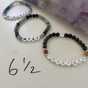May include: Three beaded bracelets with white letter beads spelling "CREATURES", "MYST", and "HONEYBEE". The bracelets are made with various colored stones and beads. The size is written as 6 1/2 inches.