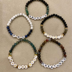 May include: Five beaded bracelets with white letter beads spelling out "Arcadia", "EMG", "Into the Myst", "Apadia", and "Creatures". The bracelets feature various colored beads, including black, green, white, and brown, with gold accents.