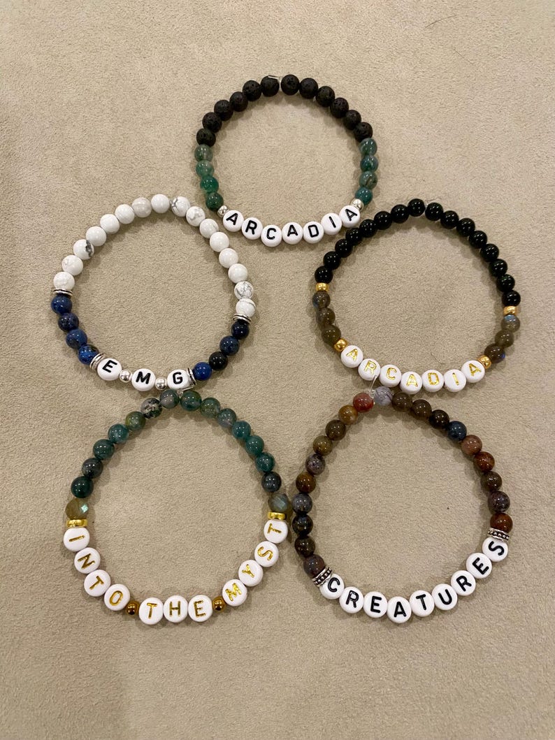 May include: Five beaded bracelets with white letter beads spelling out "Arcadia", "EMG", "Into the Myst", "Apadia", and "Creatures". The bracelets feature various colored beads, including black, green, white, and brown, with gold accents.