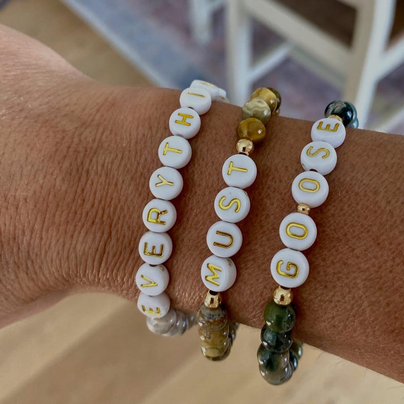 May include: Three beaded bracelets spelling out "EVERYTHING MUST GOOSE". The bracelets feature white beads with gold lettering, interspersed with gemstone beads in shades of brown, green, and gold. The bracelets are worn on a wrist.