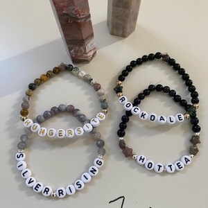 May include: Four beaded bracelets with white letter beads spelling "SILVER RISING", "HUNGERSITE", "ROCKDALE", and "HOTTEA". The bracelets feature various colored beads and gold accents. Two stone obelisks are in the background.