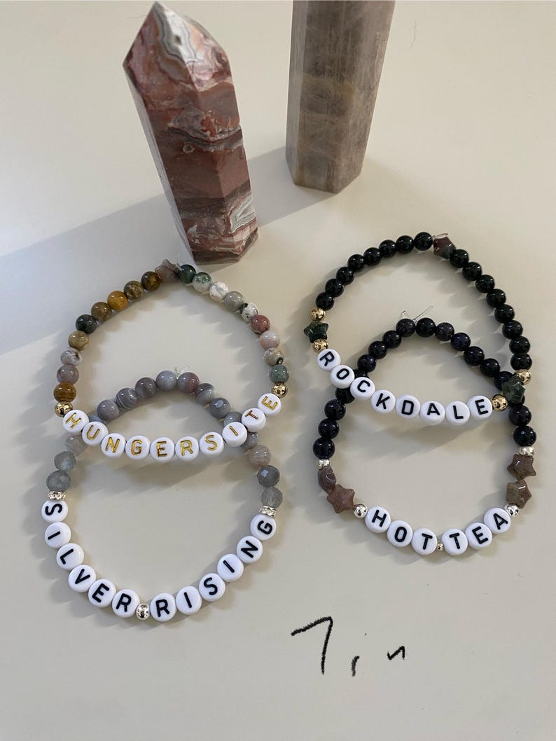 May include: Four beaded bracelets with white letter beads spelling "SILVER RISING", "HUNGERSITE", "ROCKDALE", and "HOTTEA". The bracelets feature various colored beads and gold accents. Two stone obelisks are in the background.