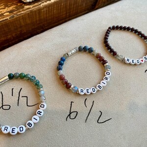 May include: Three beaded bracelets with the word "REDBIRD" spelled out in white letter beads. One bracelet is green, one is a mix of blue and red, and the third is dark red. The bracelets are approximately 6.5 inches and 7 inches in circumference.