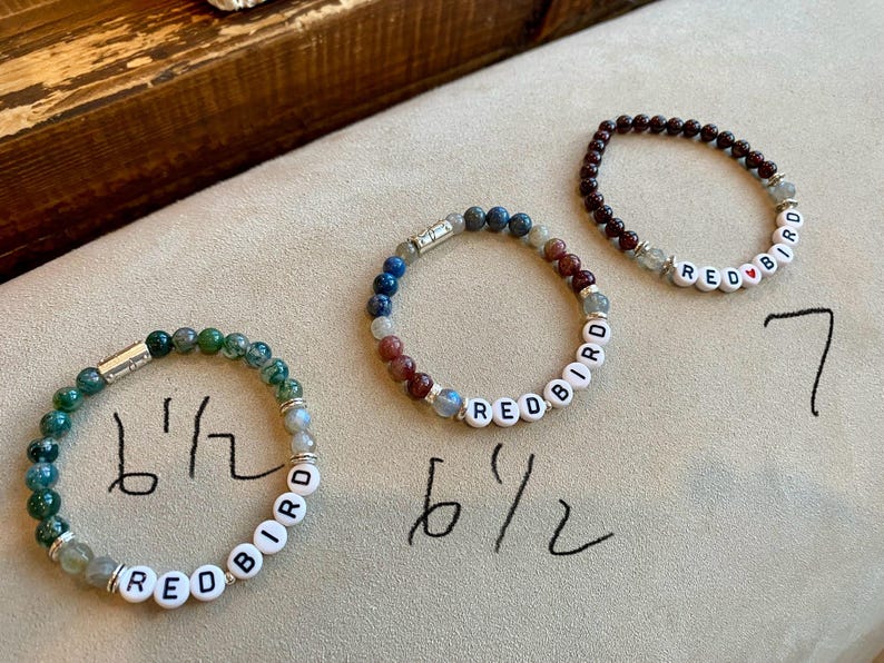 May include: Three beaded bracelets with the word "REDBIRD" spelled out in white letter beads. One bracelet is green, one is a mix of blue and red, and the third is dark red. The bracelets are approximately 6.5 inches and 7 inches in circumference.