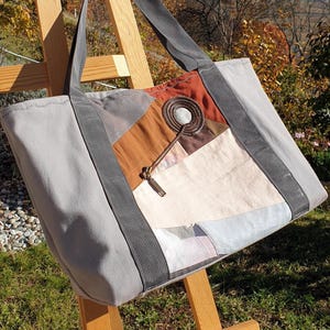 Peut inclure: Un sac fourre-tout gris avec un motif patchwork sur le devant. Le patchwork comprend des carrés de tissu marron, beige et blanc cassé. Le sac a des sangles grises et un détail de fermeture éclair. Le sac est exposé sur un chevalet en bois.