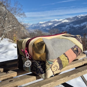 Peut inclure: Un sac rectangulaire en patchwork avec une fermeture éclair bordeaux et un charm smiley argenté. Le sac présente divers motifs de tissu dans les tons de vert, jaune, gris et rose. Le sac repose sur un banc en bois avec un fond de montagnes enneigées.