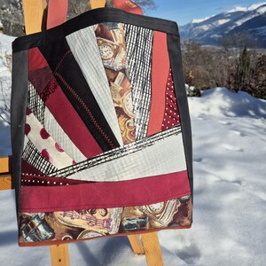 Peut inclure: Sac fourre-tout patchwork avec des tissus rouges, noirs et blancs. Le sac présente un motif avec des cadrans d'horloge et d'autres motifs. Il a des anses rouges et est posé sur un fond enneigé.