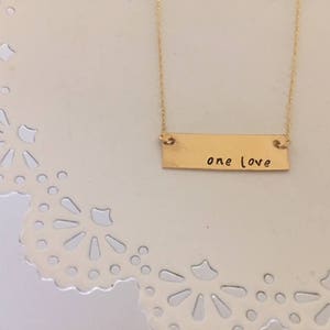 May include: A gold-colored necklace with a rectangular pendant. The pendant has the words "one love" engraved in a handwritten style. The necklace is displayed on a white, decorative surface.