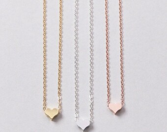 May include: Three delicate necklaces with heart pendants. The necklaces are in gold, silver, and rose gold. Each necklace features a small heart-shaped pendant suspended from a fine chain. The pendants are smooth and polished, reflecting light.