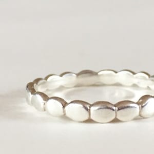 May include: A silver ring with a band of small, evenly spaced, rounded shapes. The ring is set against a white background, and the metal has a bright, reflective quality. The ring appears to be a delicate piece of jewelry.
