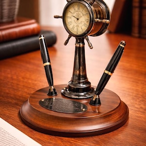 May include: A vintage-style desk clock with a ship's wheel design, featuring a clock face with Roman numerals. The clock is mounted on a wooden base with two black pens. The base also has a decorative metal plate.