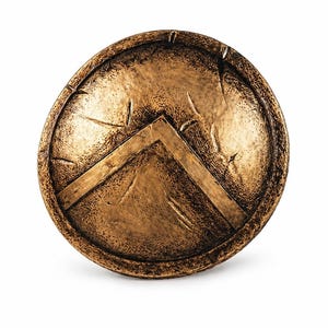 May include: A bronze-colored, round shield with a prominent chevron design. The shield has a textured surface with visible scratches and wear, suggesting an aged or battle-worn appearance. The shield is a prop or decorative item.