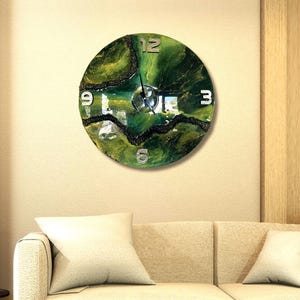 Può includere: Un orologio da parete rotondo con un design astratto verde e nero. Il quadrante dell'orologio presenta numeri argentati nelle posizioni 12, 3, 6 e 9. L'orologio è montato su una parete beige sopra un divano beige.
