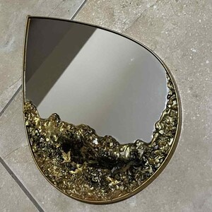 以下が含まれることがあります： 金色の金属フレームが付いた、涙の形をした鏡。 下端には、テクスチャ加工された金色の装飾が施されています。 鏡の表面は周囲の環境を映し出しています。
