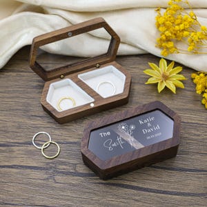 May include: Two wooden ring boxes with clear lids. One box is open, displaying two rings in white velvet compartments. The other box has a clear lid with the text "The Smith" and "Katie & David 06.03.2025".
