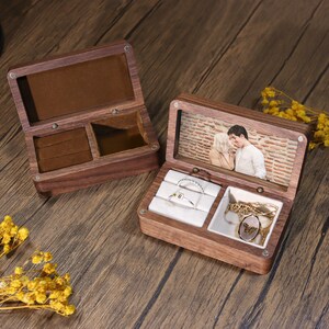 K&ouml;nnte beinhalten: Zwei offene Schmuckk&auml;stchen aus Holz. Eine Box hat einen braunen Samtinnenraum und F&auml;cher. Die andere Box hat ein Foto eines sich k&uuml;ssenden Paares unter dem Deckel, mit Ringen und einer Halskette im Inneren. Die Boxen stehen auf einer Holzoberfl&auml;che.