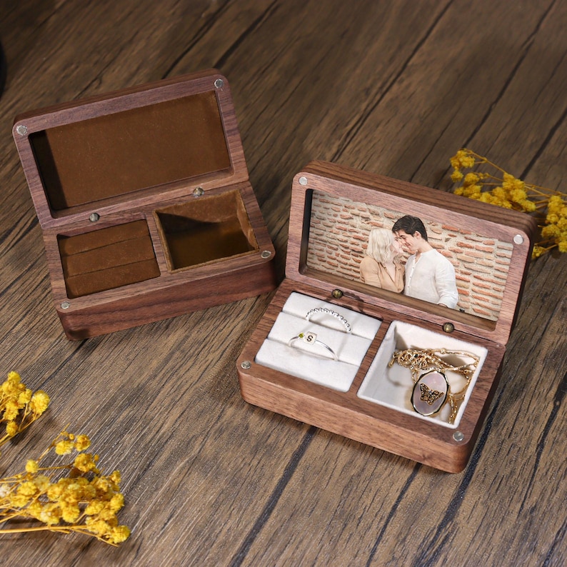K&ouml;nnte beinhalten: Zwei offene Schmuckk&auml;stchen aus Holz. Eine Box hat einen braunen Samtinnenraum und F&auml;cher. Die andere Box hat ein Foto eines sich k&uuml;ssenden Paares unter dem Deckel, mit Ringen und einer Halskette im Inneren. Die Boxen stehen auf einer Holzoberfl&auml;che.
