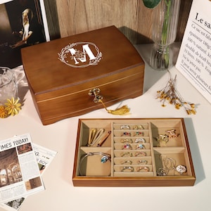 May include: A brown wooden jewelry box with a floral "M" design on the lid. The open box reveals a tray with rings, bracelets, and other jewelry. The box has a gold-colored clasp and a tassel. The tray is lined with beige fabric.