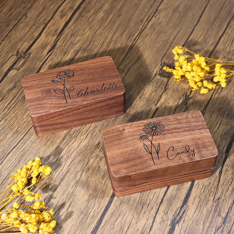 K&ouml;nnte beinhalten: Zwei rechteckige Holzkisten mit eingravierten Blumendesigns und Namen. Eine Schachtel zeigt eine Rose und den Namen "Charlotte", die andere G&auml;nsebl&uuml;mchen und den Namen "Candy". Die Kisten stehen auf einer Holzoberfl&auml;che.