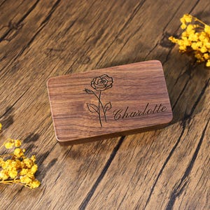 K&ouml;nnte beinhalten: Eine rechteckige Holzbox mit einer Rosen-Gravur und dem Namen "Charlotte" in Schreibschrift. Die Box ist aus dunkelbraunem Holz gefertigt und hat eine glatte Oberfl&auml;che. Die Rose ist mit detaillierten Linien dargestellt, und der Name ist elegant geschrieben.