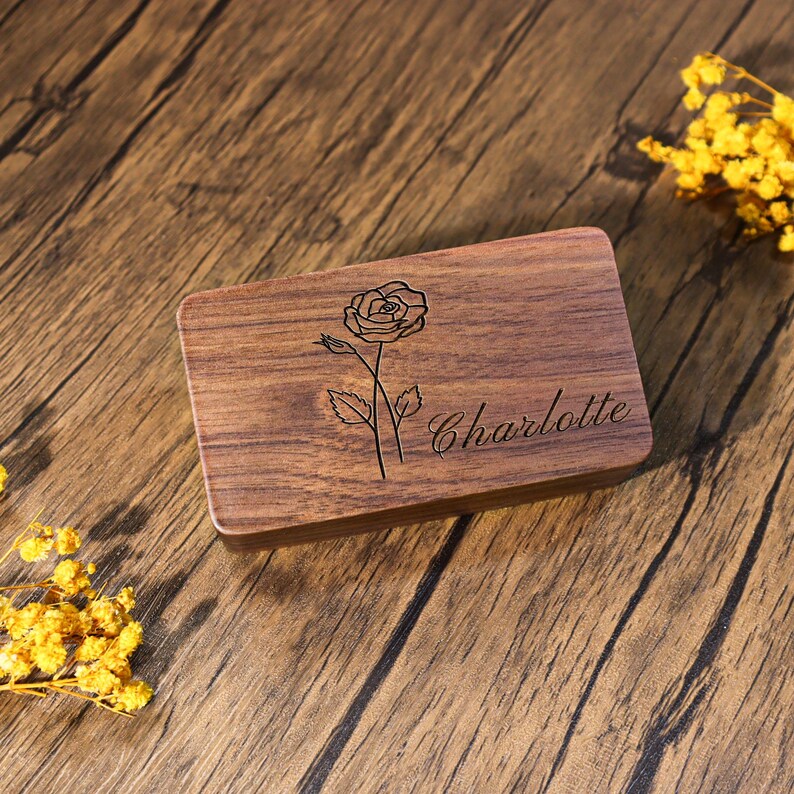 K&ouml;nnte beinhalten: Eine rechteckige Holzbox mit einer Rosen-Gravur und dem Namen "Charlotte" in Schreibschrift. Die Box ist aus dunkelbraunem Holz gefertigt und hat eine glatte Oberfl&auml;che. Die Rose ist mit detaillierten Linien dargestellt, und der Name ist elegant geschrieben.