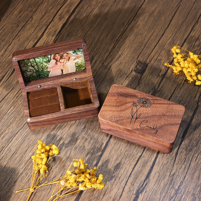K&ouml;nnte beinhalten: Ge&ouml;ffnete Holzschmuckschatulle mit Foto im Deckel und eine separate Holzbox mit Blumendesign und dem Namen "Candy". Die offene Box hat Ringschlitze und ein Samtfutter. Gelbe Blumen sind verstreut.