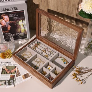 Peut inclure: Une boîte à bijoux en bois avec un couvercle en verre, remplie de bagues, colliers et bracelets. La boîte ouverte révèle plusieurs compartiments doublés de tissu gris. Le nom "Emily" est gravé sur le devant.