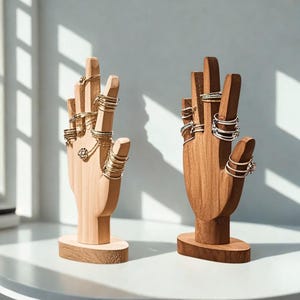 May include: Two wooden hand-shaped ring holders. One is light-colored, the other is dark. Both hold multiple rings of various styles and colors, including gold and silver. The holders sit on a white surface.