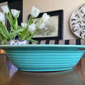 May include: A turquoise ceramic bowl with horizontal ridged detailing. The bowl is on a wooden surface, with a vase of white tulips in the background. A decorative plate and framed artwork are also visible.