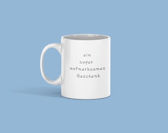 May include: White ceramic mug with a curved handle, featuring the German text "ein super aufmerksames Geschenk" in black, centered on the front. The mug is set against a light blue background.