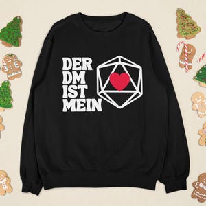 Puede incluir: Sudadera negra con el texto "DER DM IST MEIN" y una forma geométrica blanca con un corazón rojo. La imagen está decorada con galletas navideñas, hombres de jengibre, bastones de caramelo y árboles de Navidad.