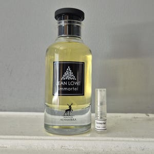 May include: A clear glass perfume bottle with a black cap and a small sample vial. The bottle has a black label with white text that reads "JEAN LOWE immortel" and a deer logo. The perfume is a light yellow color.
