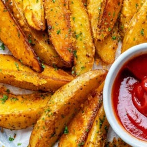 May include: Close-up of golden-brown potato wedges seasoned with herbs. A small white bowl filled with bright red ketchup is in the corner. The wedges are crispy and appear freshly cooked, perfect for a snack or side dish.