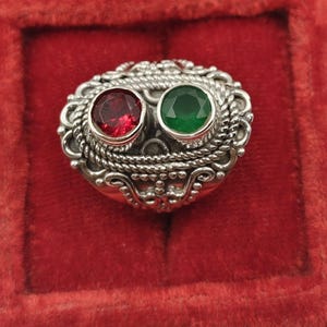 May include: Ornate silver ring featuring two gemstones: a round, deep red stone and a round, vibrant green stone. The ring has a detailed, textured design with a rope-like border and decorative flourishes, set against a red velvet background.