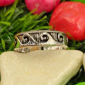Puede incluir: Un anillo de plata con un diseño de olas ornamentado. El anillo presenta un patrón detallado de líneas onduladas y barras verticales. El anillo se muestra sobre un fondo neutro con acentos verdes y rojos.