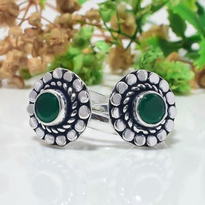 May include: A silver ring featuring two circular settings, each containing a smooth, round, dark green stone. The settings are surrounded by a decorative border of small silver spheres and a twisted silver wire detail, creating a floral appearance.