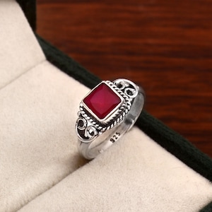 Op de afbeelding: Een zilveren ring met een vierkante, dieprode edelsteen. De ring heeft een sierlijk ontwerp met een gedraaide touwrand rond de steen en decoratieve krullen aan de zijkanten. Het nummer "925" is op de band gestempeld.