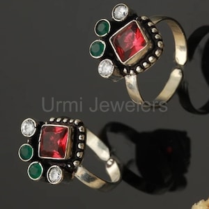 May include: Two silver toe rings with a square red gemstone center, surrounded by a beaded border. Each ring has two green and two white gemstones. The rings are displayed on a reflective black surface.