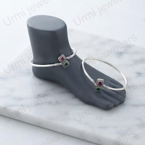 May include: A pair of silver anklets featuring a thin, smooth band and a central decorative element composed of two square-shaped settings. Each setting contains a small, inlaid gemstone, one in a deep red and the other in a vibrant green.
