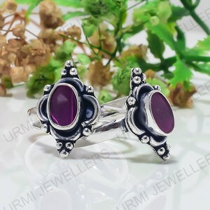 May include: Two silver rings featuring an oval-shaped deep magenta stone set in a decorative metal frame with small silver spheres. The metal has a polished finish with darkened recessed areas. The text Urmi Jewellers is visible in the background.