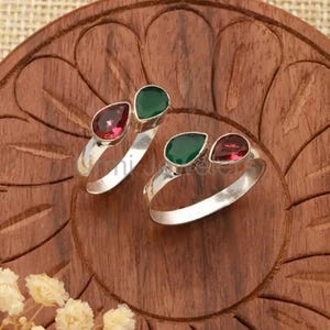 May include: Two silver toe rings, each featuring a teardrop-shaped green and red gemstone. The rings are displayed on a carved wooden surface, showcasing the jewelry's design and color contrast.