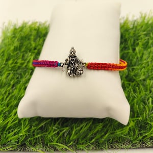 May include: A colorful woven bracelet with a silver charm of a deity. The bracelet features a braided design with red, pink, blue, and yellow threads. The charm is a detailed depiction of a seated figure holding objects. The bracelet is displayed on a white pillow.