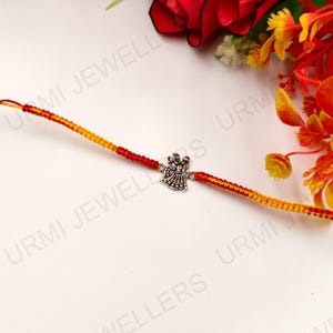 May include: A decorative bracelet featuring a central silver-toned metal charm depicting two figures in traditional attire. The band is crafted from braided orange and yellow thread, creating a vibrant, textured cord that extends from the charm.