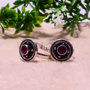 Può includere: Un paio di anelli color argento con una faccia circolare e una pietra centrale color magenta intenso. L'incastonatura circolare è accentuata da un bordo a corda intrecciata e piccoli punti argentati. La fascia è aperta e regolabile.