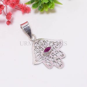May include: Silver Hamsa hand pendant with detailed openwork. It has a central oval, deep pink gemstone and the text "URMA RS". The pendant hangs from a silver bail.