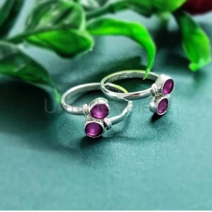 May include: Two silver toe rings, each featuring two oval, magenta-colored gemstones. The rings are displayed on a teal surface, with green foliage in the background. The gemstones are set in a simple, elegant design.
