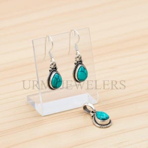 May include: A silver jewelry set featuring a pair of teardrop earrings and a matching pendant. Each piece has a turquoise stone set in a decorative silver frame. The earrings hang from silver hooks.