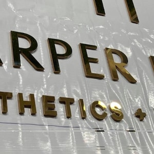 May include: Gold metallic letters spelling "HARPER HAUS AESTHETICS + WELLNESS" on a white surface. The letters are three-dimensional and reflect light, creating a shiny appearance. The sign is likely for a business.