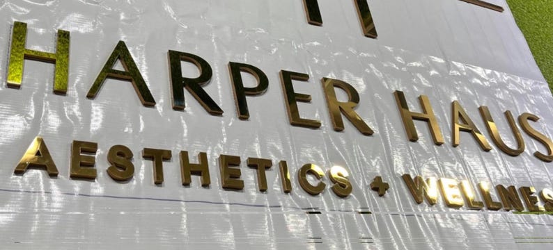 May include: Gold metallic letters spelling "HARPER HAUS AESTHETICS + WELLNESS" on a white surface. The letters are three-dimensional and reflect light, creating a shiny appearance. The sign is likely for a business.